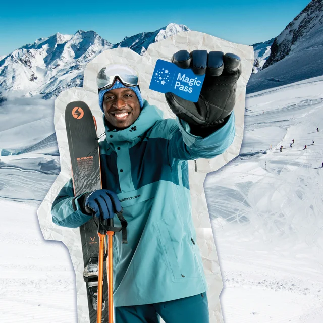 Un skieur souriant, habillé en tenue de ski bleue, tenant un forfait Magic Pass dans une main gantée et des skis dans l'autre, devant un paysage enneigé de haute montagne.