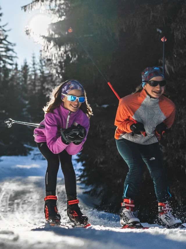 Trois skieurs de fond, souriants et équipés, glissent sur une piste enneigée à travers une forêt de sapins sous le soleil d'hiver.