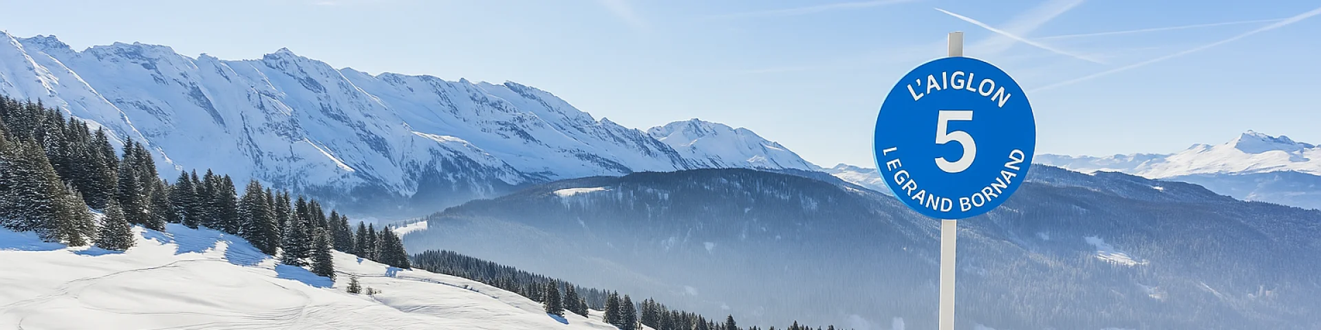 Panneau bleu indiquant la piste L'Aiglon numéro 5 à la station de ski Le Grand Bornand, avec un paysage enneigé et des montagnes en arrière-plan.