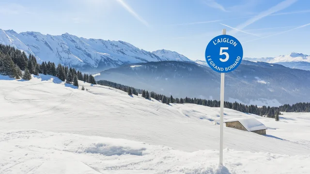Panneau bleu indiquant la piste L'Aiglon numéro 5 à la station de ski Le Grand Bornand, avec un paysage enneigé et des montagnes en arrière-plan.