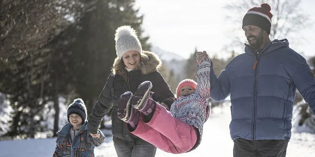 Balade à pied en hiver en famille. Le papa et la maman soulève d'une main chacun leur petite fille qui rigole et la maman tient par la main son fils.