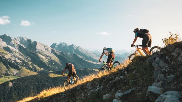 VTT sportif au Grand-Bornand. 3 personnes dévalent une pente en VTT avec pour toile de fond, la chaîne des Aravis.