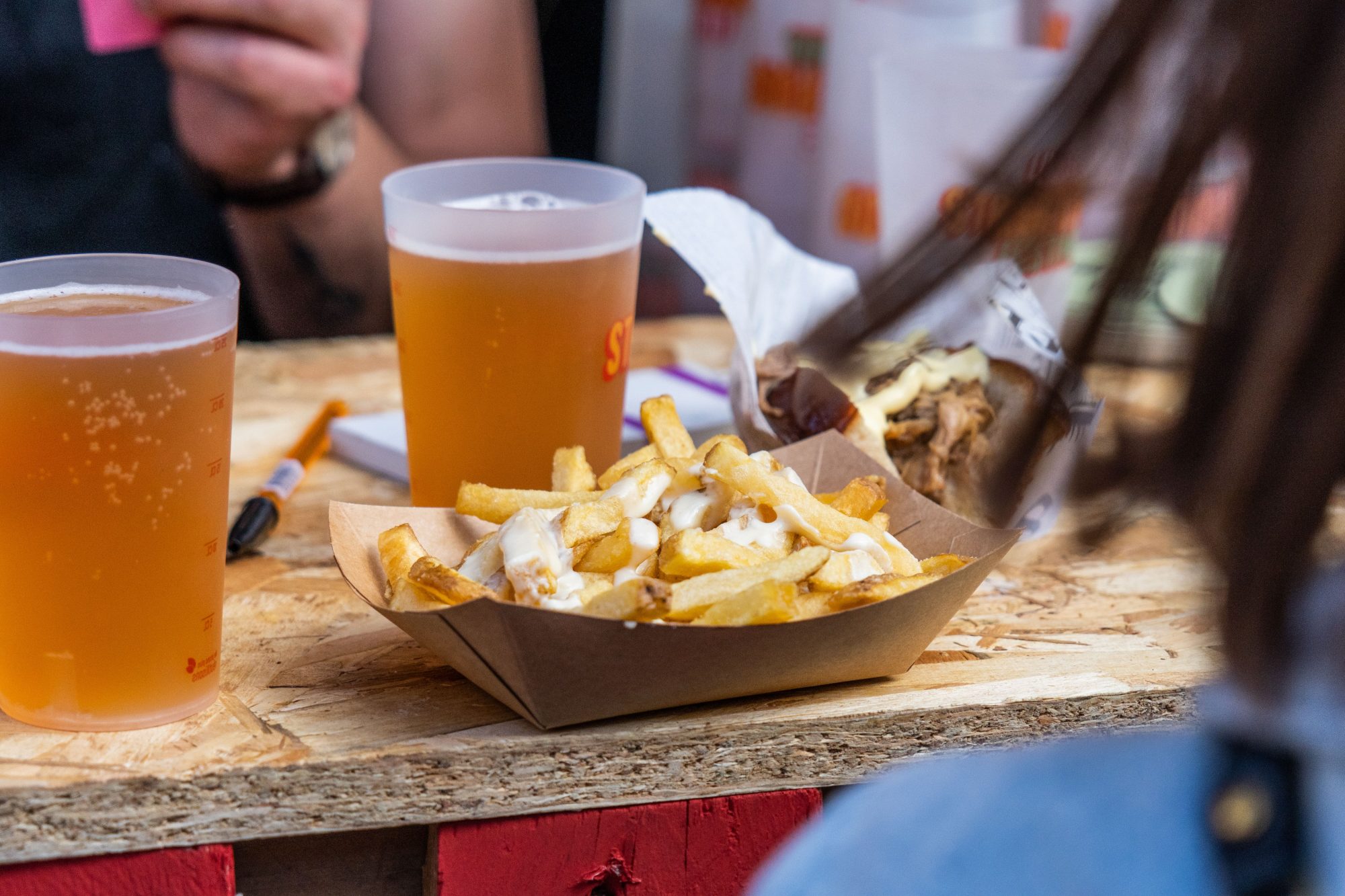 Portion de frites nappées de sauce servie dans une barquette, accompagnée de deux bières blondes lors d’un événement gastronomique.