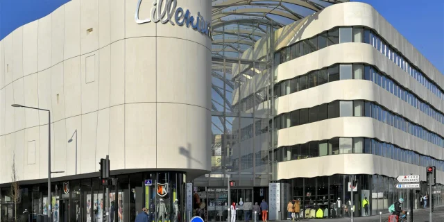 Façade du centre commercial Lillenium à Lille, reconnaissable à son architecture aux courbes ondulées et à ses murs clairs. Le nom Lillenium est affiché en lettres métalliques sur la partie gauche. Le centre comporte une entrée vitrée sous une grande verrière incurvée. Des vitrines de magasins sont visibles au rez-de-chaussée. Plusieurs piétons et un cycliste en train de traverser sont présents au premier plan. Le ciel est dégagé et bleu.