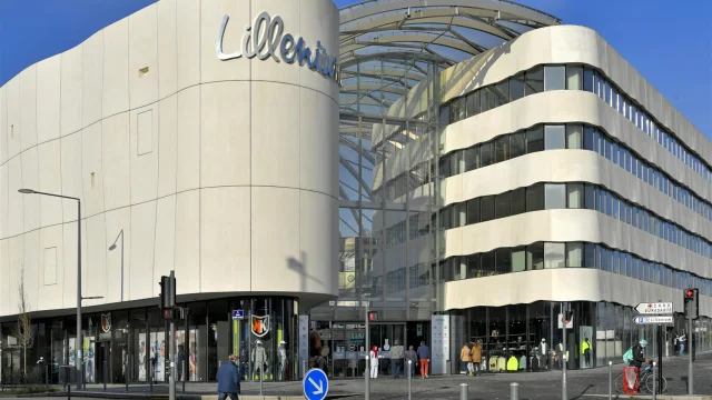 Façade du centre commercial Lillenium à Lille, reconnaissable à son architecture aux courbes ondulées et à ses murs clairs. Le nom Lillenium est affiché en lettres métalliques sur la partie gauche. Le centre comporte une entrée vitrée sous une grande verrière incurvée. Des vitrines de magasins sont visibles au rez-de-chaussée. Plusieurs piétons et un cycliste en train de traverser sont présents au premier plan. Le ciel est dégagé et bleu.