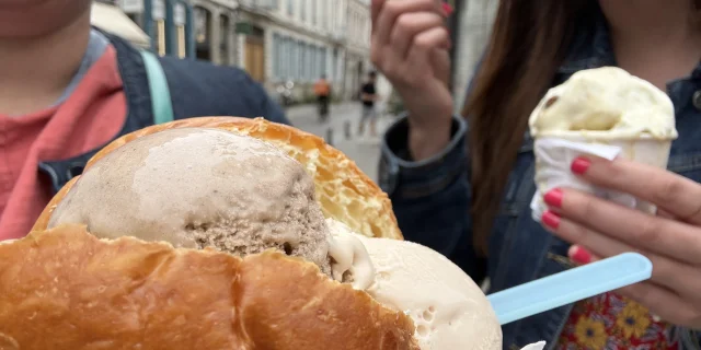 Deux personnes dégustent des glaces dans une rue piétonne bordée d'immeubles anciens. Au premier plan, une glace est servie dans une brioche moelleuse, avec deux boules (une beige et une marron clair), accompagnée d’une cuillère en plastique bleue. Elle est tenue dans un papier blanc. En arrière-plan, une autre personne tient un petit pot de glace vanille avec également une cuillère bleue. Cette personne porte du vernis rose vif sur les ongles et une veste en jean sur une robe à fleurs colorée. La première personne, partiellement visible, porte un haut corail et un sac turquoise.