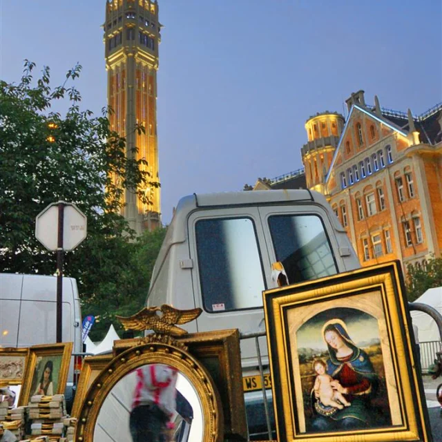 Scène de la Braderie de Lille en soirée, sous un ciel bleu crépusculaire. Au premier plan, un étal de brocante exposant des cadres dorés, un miroir rond reflétant un passant flou, et plusieurs tableaux, dont une représentation de la Vierge à l’Enfant. En arrière-plan, des camionnettes blanches et des bâtiments de style flamand, dont la tour de l’Hôtel de Ville de Lille illuminée en jaune, dominant la scène. L’ambiance est à la fois animée et pittoresque.