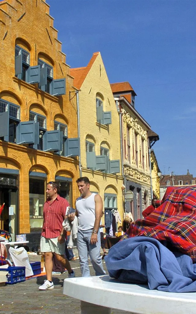 Scène de braderie sous un ciel bleu à Lille, avec des passants marchant le long de bâtiments anciens en briques colorées. Au premier plan, un tas de vêtements d’occasion est posé sur une table, tandis qu’un enfant regarde le sol en portant un sac noir. En arrière-plan, plusieurs personnes déambulent entre les étals installés dans la rue pavée.