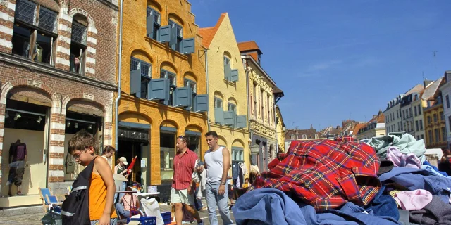 Scène de braderie sous un ciel bleu à Lille, avec des passants marchant le long de bâtiments anciens en briques colorées. Au premier plan, un tas de vêtements d’occasion est posé sur une table, tandis qu’un enfant regarde le sol en portant un sac noir. En arrière-plan, plusieurs personnes déambulent entre les étals installés dans la rue pavée.