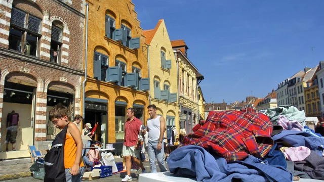 Scène de braderie sous un ciel bleu à Lille, avec des passants marchant le long de bâtiments anciens en briques colorées. Au premier plan, un tas de vêtements d’occasion est posé sur une table, tandis qu’un enfant regarde le sol en portant un sac noir. En arrière-plan, plusieurs personnes déambulent entre les étals installés dans la rue pavée.