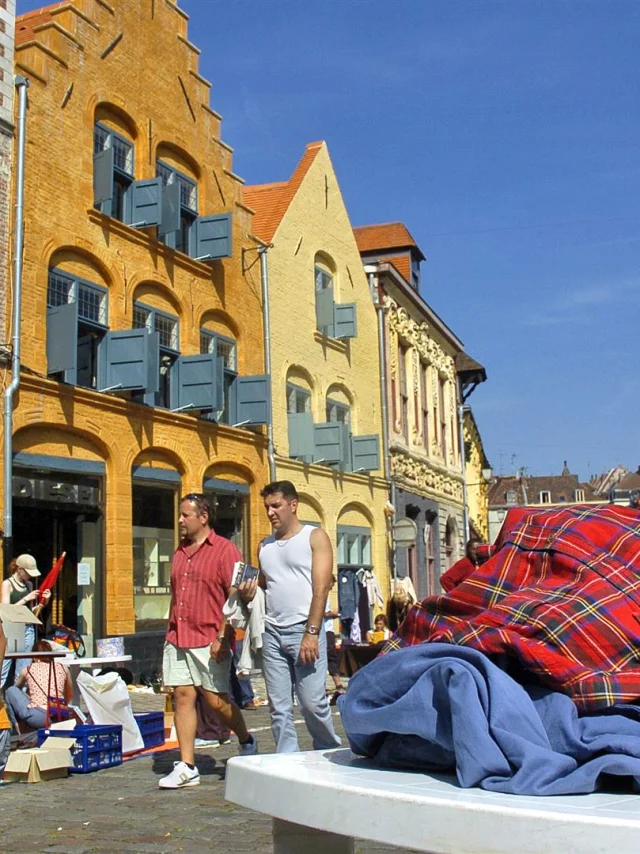 Scène de braderie sous un ciel bleu à Lille, avec des passants marchant le long de bâtiments anciens en briques colorées. Au premier plan, un tas de vêtements d’occasion est posé sur une table, tandis qu’un enfant regarde le sol en portant un sac noir. En arrière-plan, plusieurs personnes déambulent entre les étals installés dans la rue pavée.
