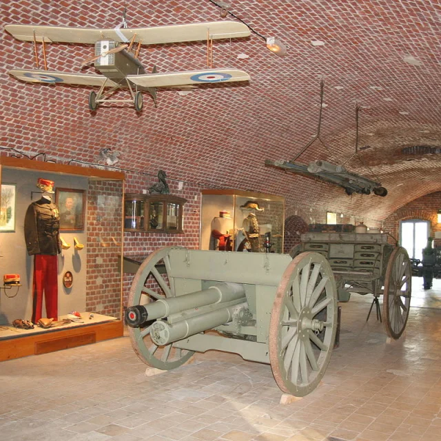Salle d’exposition du Fort de Seclin présentant des pièces militaires, dont un canon, un uniforme et une maquette d’avion suspendue.