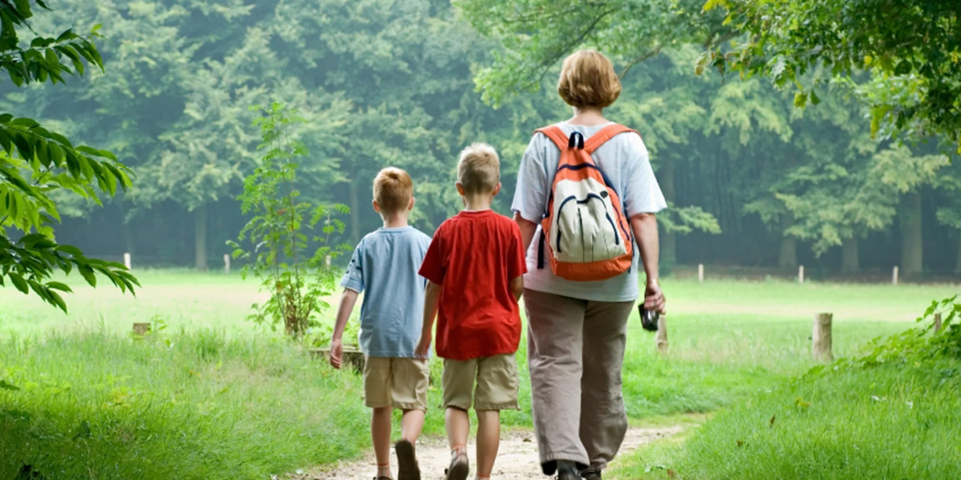 Une femme et deux enfants marchent sur un sentier dans la nature, entourés de verdure, en direction d’un bois .