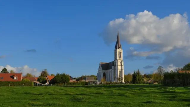 Vue lointaine de l’église de Bouvines au milieu de prairies verdoyantes et de maisons aux toits rouges.