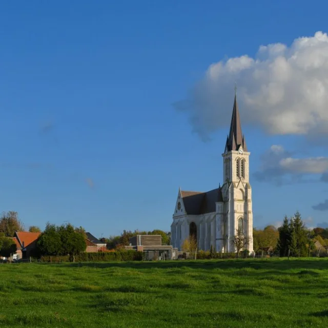 Vue lointaine de l’église de Bouvines au milieu de prairies verdoyantes et de maisons aux toits rouges.