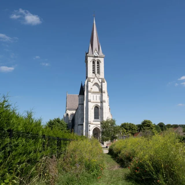 Chemin bordé de hautes herbes menant à l’église de Bouvines sous un ciel bleu.