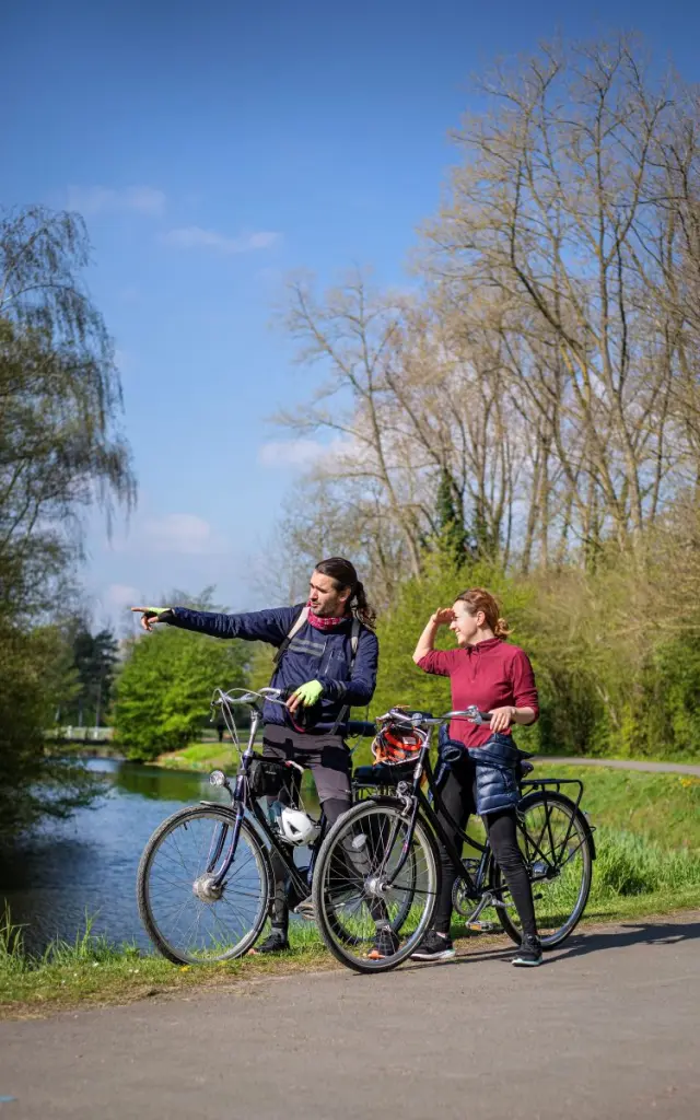 Un couple à vélo s’arrêtant près d’un canal, l’homme montrant un point au loin.
