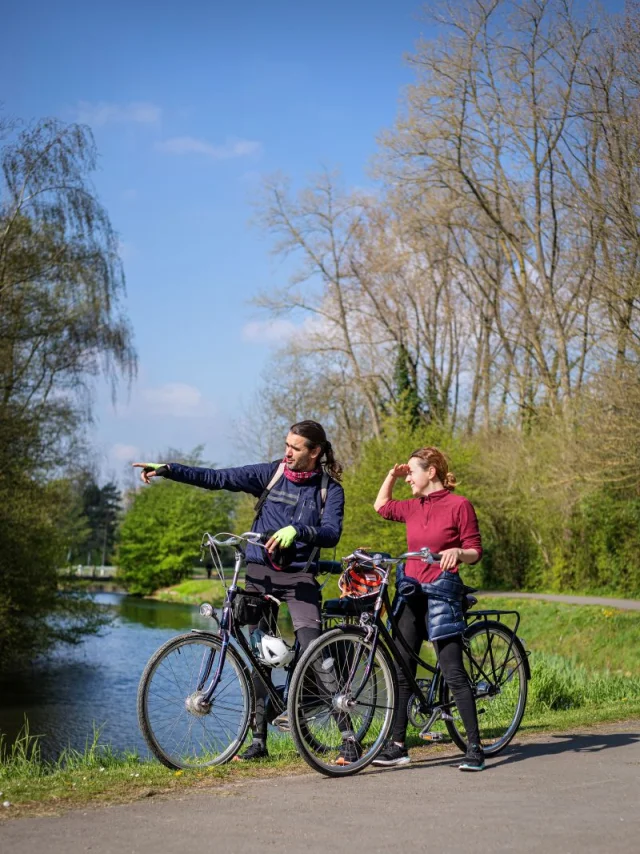 Un couple à vélo s’arrêtant près d’un canal, l’homme montrant un point au loin.