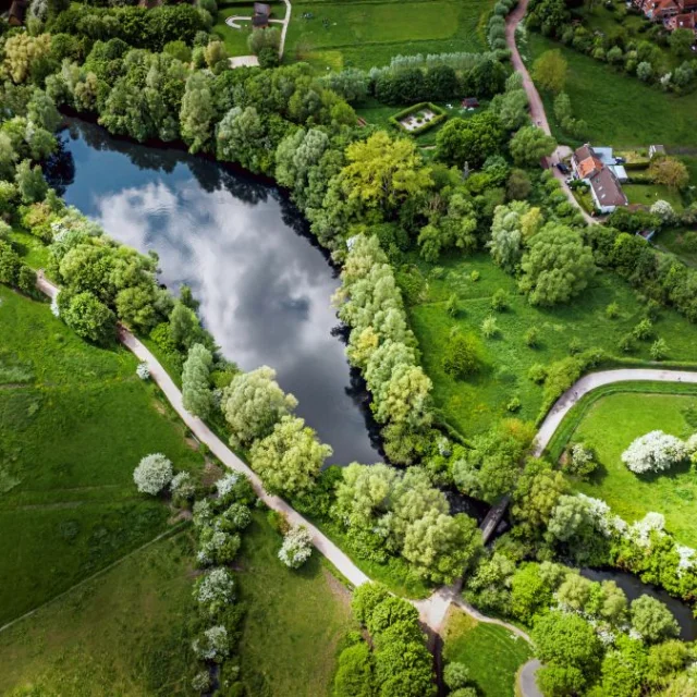 Vue aérienne du Lac du Héron entouré d’arbres et de sentiers, près de maisons rurales dans un paysage verdoyant.