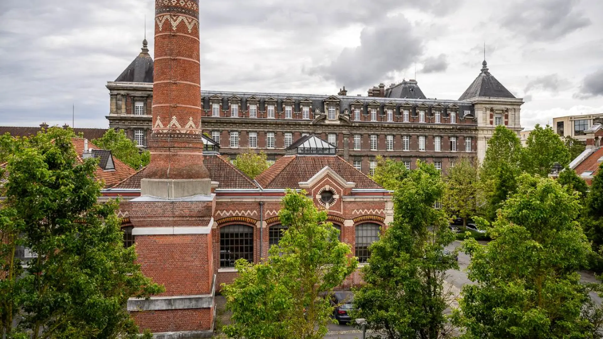 Cheminée en brique rouge ornée de motifs géométriques devant les bâtiments historiques de l'École nationale des arts et métiers de Lille, entourée d’arbres et sous un ciel nuageux.