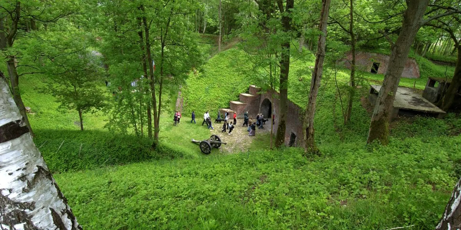 Entrée du Fort de Seclin vue depuis un talus boisé, avec un groupe de visiteurs et un canon ancien installé sur la pelouse.