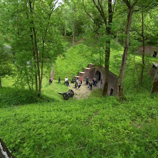 Entrée du Fort de Seclin vue depuis un talus boisé, avec un groupe de visiteurs et un canon ancien installé sur la pelouse.