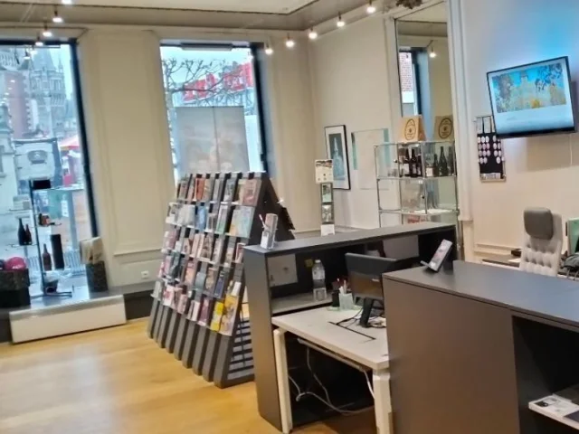 Espace d’accueil de l’Office de Tourisme de Tourcoing avec comptoirs modernes, présentoirs de brochures et vue sur la Grand-Place par les fenêtres.