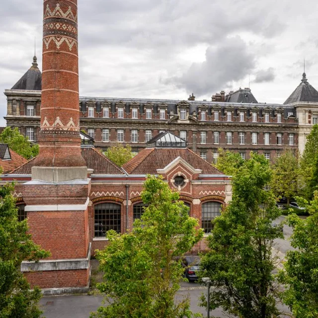 Cheminée en brique rouge ornée de motifs géométriques devant les bâtiments historiques de l'École nationale des arts et métiers de Lille, entourée d’arbres et sous un ciel nuageux.