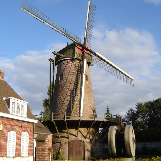 Moulin en brique de style traditionnel avec ailes à claire-voie, situé à Halluin, entouré de fleurs et de meules de pierre exposées.