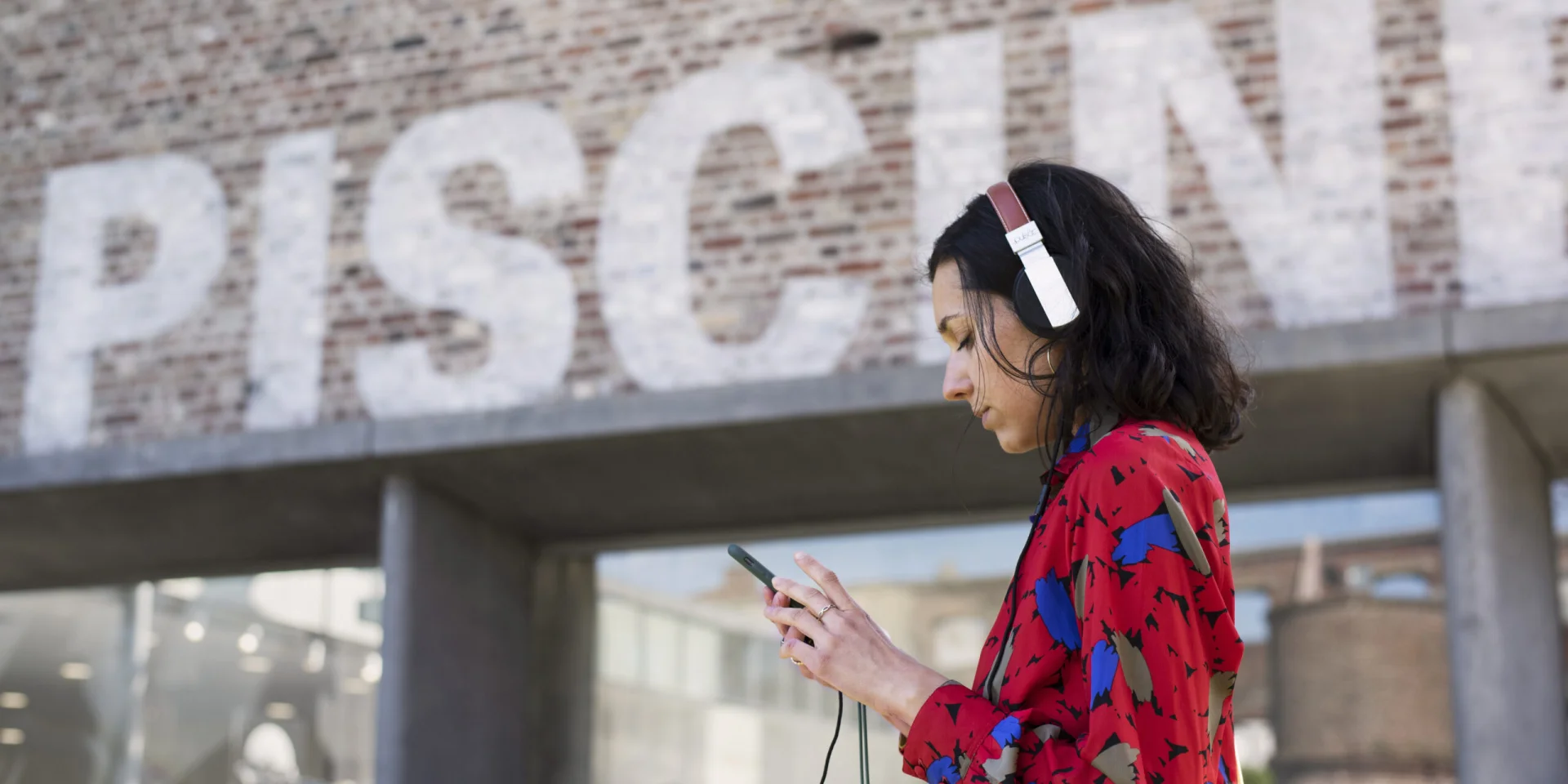 Femme avec un casque audio et un smartphone devant l'entrée en briques de La Piscine, musée d'art et d'industrie à Roubaix.