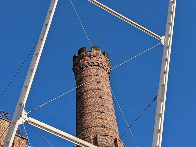 Cheminée en briques du site Motte-Bossut à Roubaix, vue à travers une structure métallique sous un ciel bleu.