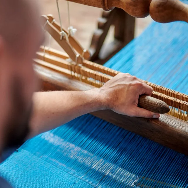 Main d’un artisan actionnant un métier à tisser traditionnel avec des fils bleus tendus en cours de tissage.