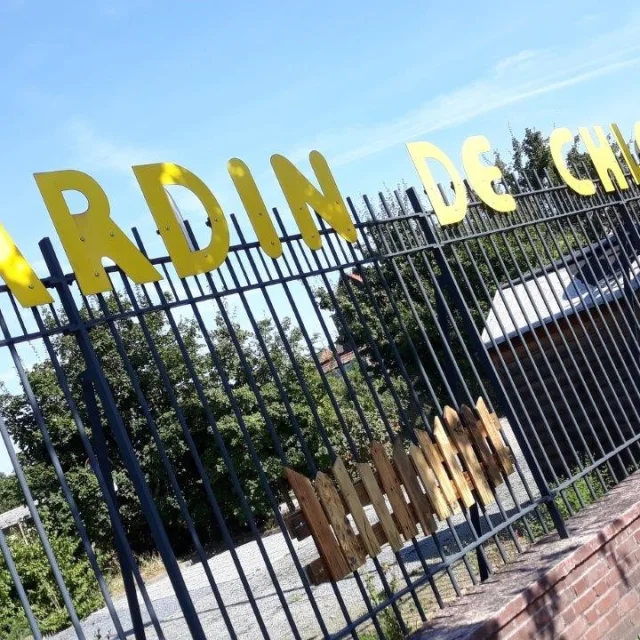 Grille noire surmontée de lettres jaunes formant “Jardin de Chlorophylle” à Roubaix, avec arbres en arrière-plan sous ciel bleu.
