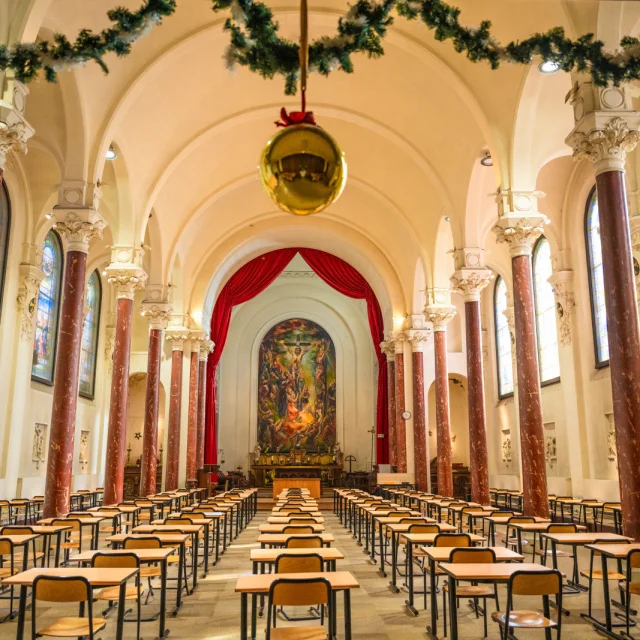 Ancienne chapelle du lycée Saint-Rémi à Roubaix, aménagée en salle d’examen, avec colonnes rouges, vitraux et fresque religieuse au fond.