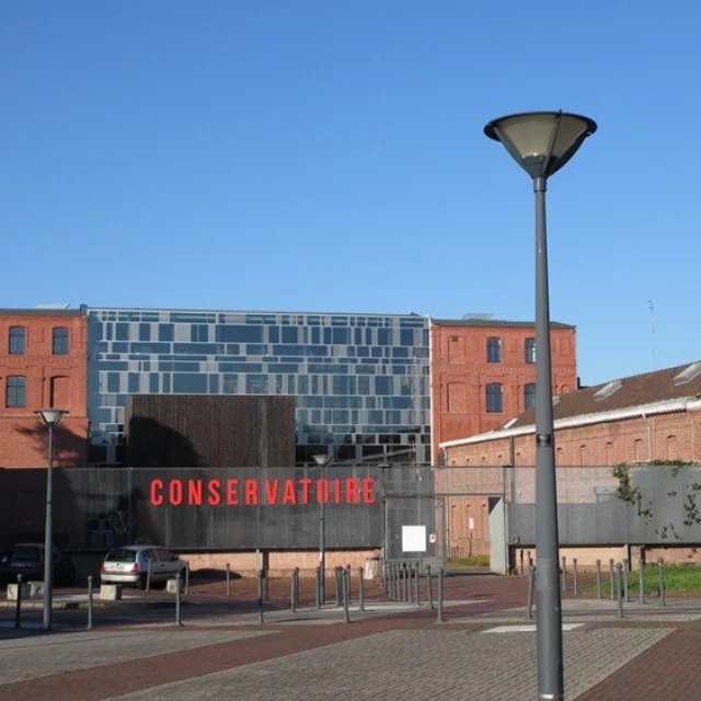 Façade du conservatoire de Roubaix, bâtiment mêlant briques rouges et structure vitrée contemporaine, sous un ciel bleu dégagé.