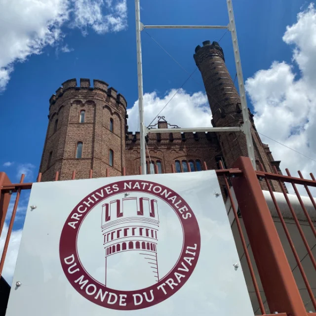 Vue en contre-plongée des Archives nationales du monde du travail à Roubaix, tours en brique rouge derrière une grille rouge et un panneau signalétique.