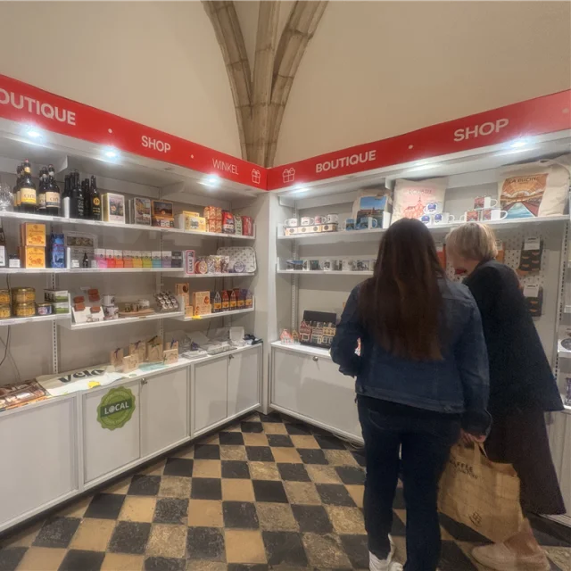 La photo montre l’intérieur d’une boutique touristique, avec des rayonnages garnis de produits locaux et souvenirs de Lille et de la Métropole : mugs, cartes postales, spécialités régionales, magnets, objets à l’effigie de la Braderie de Lille, etc. L’ambiance est lumineuse et accueillante.