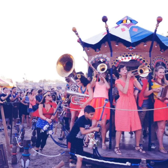 Un groupe de musiciens en fanfare, vêtus de tenues colorées, joue sur une petite scène décorée façon manège, entouré d’un public festif en plein air.