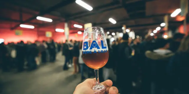 Verre de bière à la main avec le logo « BAL Bière à Lille », tenu devant une foule lors d’un événement brassicole en intérieur.