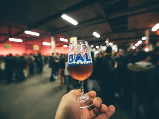 Verre de bière à la main avec le logo « BAL Bière à Lille », tenu devant une foule lors d’un événement brassicole en intérieur.
