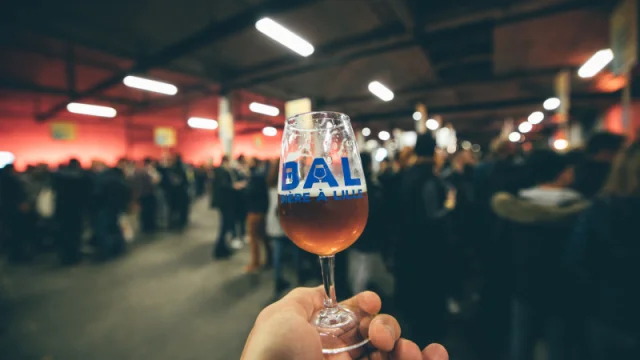 Verre de bière à la main avec le logo « BAL Bière à Lille », tenu devant une foule lors d’un événement brassicole en intérieur.