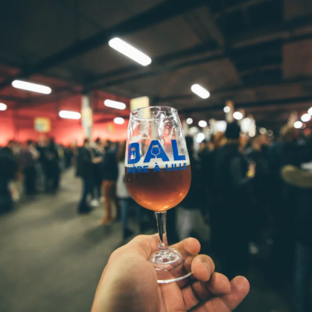 Verre de bière à la main avec le logo « BAL Bière à Lille », tenu devant une foule lors d’un événement brassicole en intérieur.