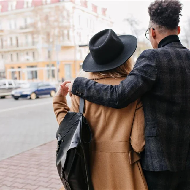 Buitenportret van afrikaanse man in geruit pak die blonde vriendin zachtjes omhelst. Meisje in beige jas geniet van uitzicht op stad tijdens date. S