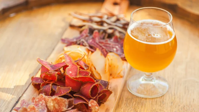 Verre de bière blonde accompagné d’une planche de charcuterie artisanale, posés sur une table en bois en extérieur.