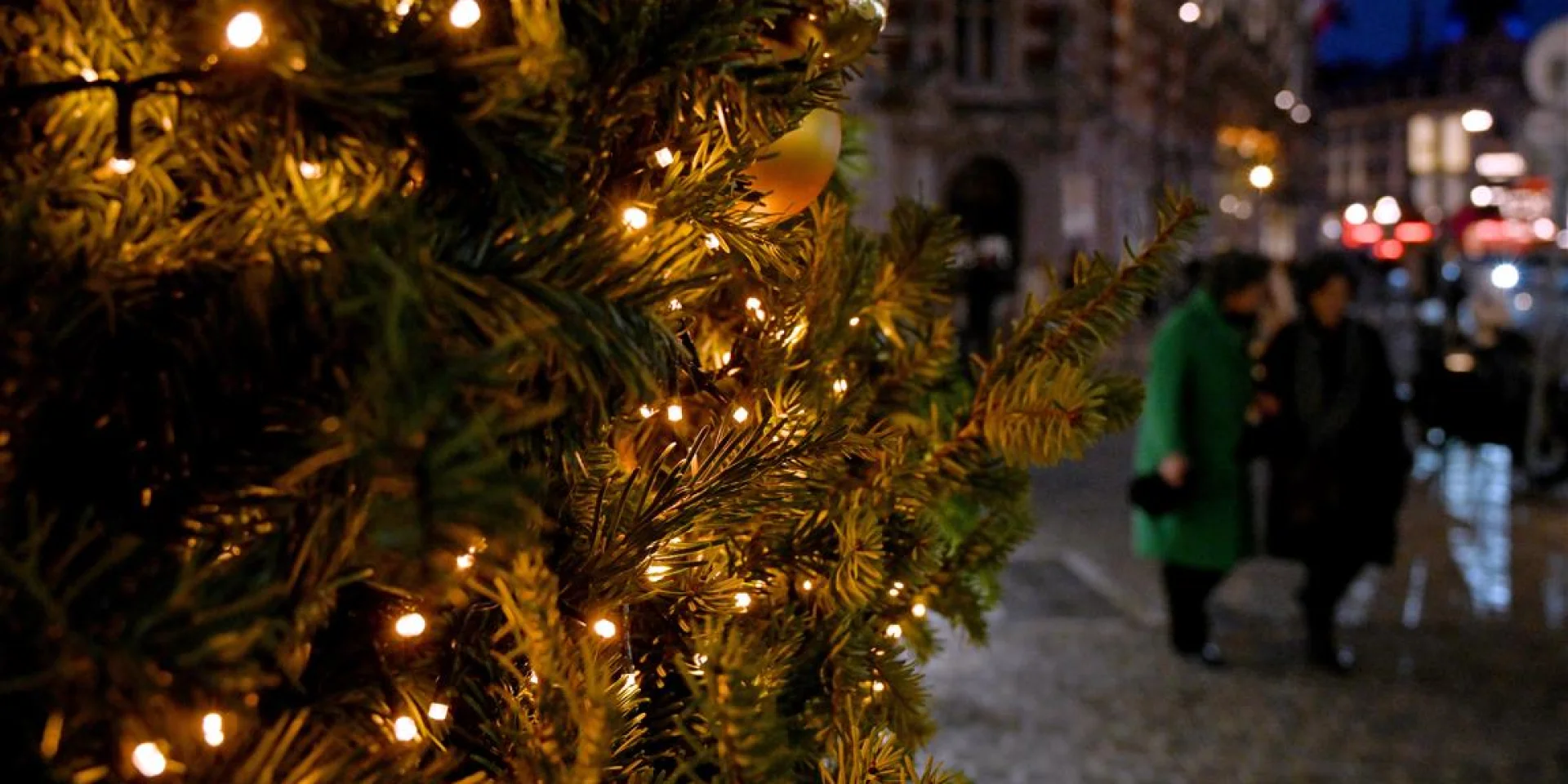 Sapin décoré de guirlandes lumineuses au premier plan, ambiance nocturne en centre-ville.