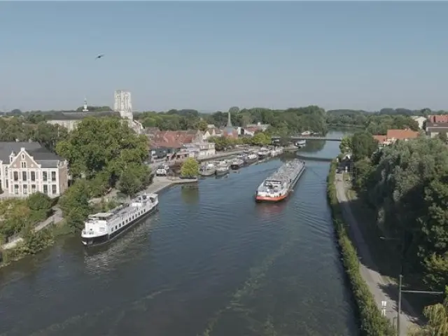 Vue aérienne du port de Wambrechies, avec ses péniches amarrées sur la Deûle, bordée de maisons et d’un château sous un ciel clair.