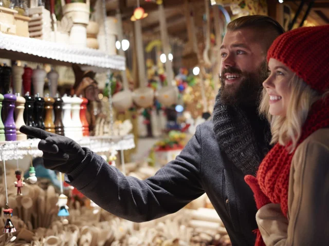 Couple souriant découvrant un stand d’artisanat coloré sur un marché de Noël, entouré de décorations et d’objets en bois.