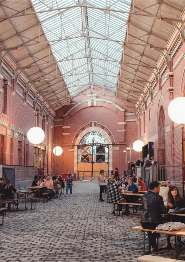 La Rue couverte de la Condition Publique à Roubaix, animée par des visiteurs discutant autour de grandes tables sous la verrière.