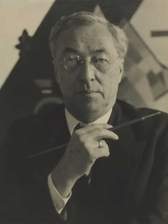 Portrait photographique en noir et blanc d’un homme âgé (peintre Kandinsky) en costume, tenant un pinceau, regard sérieux, dans un atelier d’artiste.