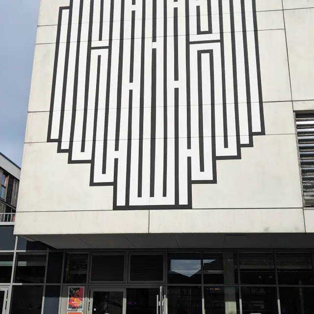 Fresque géométrique en noir et blanc de l’artiste Atlas sur la façade d’un bâtiment, composée de lignes verticales formant un motif labyrinthique.